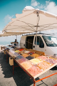 A roadside market stall features a variety of colorful dried fruits displayed in trays. The stall is set up next to a white van and shaded by a large umbrella. The background showcases a clear blue sky with a few clouds, suggesting a sunny day.