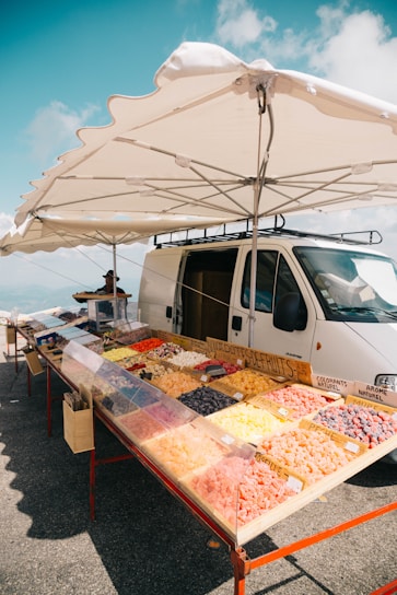 A roadside market stall features a variety of colorful dried fruits displayed in trays. The stall is set up next to a white van and shaded by a large umbrella. The background showcases a clear blue sky with a few clouds, suggesting a sunny day.