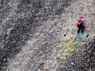 A climber is navigating a steep, textured rock face while equipped with climbing gear. They have a red helmet and backpack, and are handling ropes that are tangled and spread across the rocky surface.