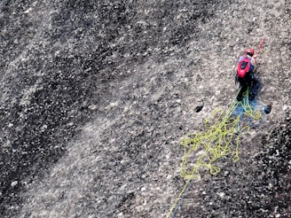 A climber is navigating a steep, textured rock face while equipped with climbing gear. They have a red helmet and backpack, and are handling ropes that are tangled and spread across the rocky surface.