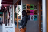 A person stands outside a building with a glass door, looking at a wall covered with colorful posters advertising various music events. The sidewalk also features a white sign for an open mic event. In the background, elements of an urban street are visible.