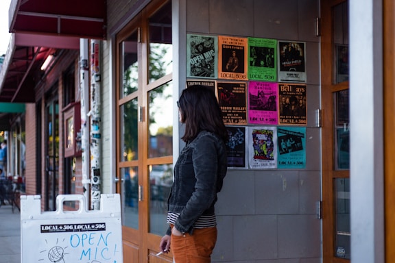 A person stands outside a building with a glass door, looking at a wall covered with colorful posters advertising various music events. The sidewalk also features a white sign for an open mic event. In the background, elements of an urban street are visible.