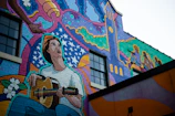 A vibrant mural painted on the side of a building features a person playing a guitar. The artwork includes swirling patterns, stars, and a stylized clock tower. In the background, silhouettes of seated individuals are visible.