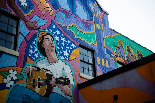 A vibrant mural painted on the side of a building features a person playing a guitar. The artwork includes swirling patterns, stars, and a stylized clock tower. In the background, silhouettes of seated individuals are visible.