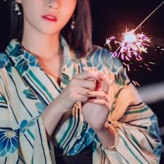 A person dressed in a floral-patterned traditional outfit holds a lit sparkler, creating a burst of colorful sparks against a dark background. The light from the sparkler illuminates their hands and part of their face.