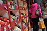 A bustling market stall filled with a variety of fresh produce including tomatoes, pears, bananas, and more, arranged on red wooden shelves. Colorful price tags are visible, and a person in a pink jacket carries a grocery bag while another holds a box.