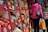 A bustling market stall filled with a variety of fresh produce including tomatoes, pears, bananas, and more, arranged on red wooden shelves. Colorful price tags are visible, and a person in a pink jacket carries a grocery bag while another holds a box.