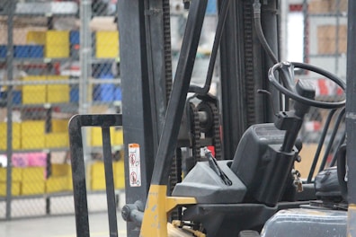 A close-up view of a forklift in an industrial warehouse setting with a focus on its mechanical parts, including the steering wheel and chain assembly. The background includes shelves filled with colorful storage bins.