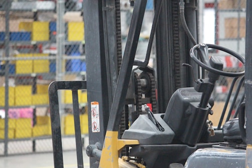 A close-up view of a forklift in an industrial warehouse setting with a focus on its mechanical parts, including the steering wheel and chain assembly. The background includes shelves filled with colorful storage bins.