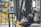 A close-up view of a forklift in an industrial warehouse setting with a focus on its mechanical parts, including the steering wheel and chain assembly. The background includes shelves filled with colorful storage bins.