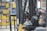 A close-up view of a forklift in an industrial warehouse setting with a focus on its mechanical parts, including the steering wheel and chain assembly. The background includes shelves filled with colorful storage bins.