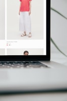 A close-up of a laptop displaying a shopping website. The screen shows a model wearing a red top and white wide-leg pants along with white sandals. The text indicates details of a round-neck t-shirt with prices mentioned.
