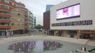 An urban square features a modern building with a large screen displaying an advertisement. The square has a decorative fountain with many small, colorful jets of water. The surroundings include a curved residential building and a few shops.