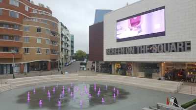 An urban square features a modern building with a large screen displaying an advertisement. The square has a decorative fountain with many small, colorful jets of water. The surroundings include a curved residential building and a few shops.