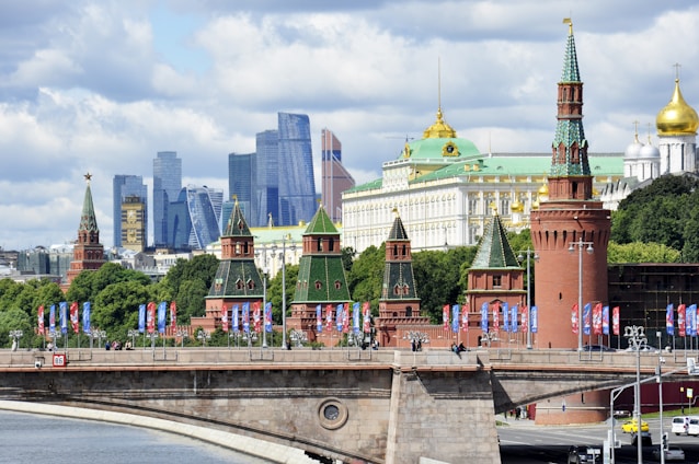 A view of iconic Russian architecture along a river, with tall modern skyscrapers in the background. The foreground features a historic red brick wall with towers topped by green spires, adorned with decorations. Flags and pedestrians are visible on a bridge that spans across the river, adding a lively atmosphere to the scene.