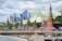 A view of iconic Russian architecture along a river, with tall modern skyscrapers in the background. The foreground features a historic red brick wall with towers topped by green spires, adorned with decorations. Flags and pedestrians are visible on a bridge that spans across the river, adding a lively atmosphere to the scene.