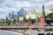 A view of iconic Russian architecture along a river, with tall modern skyscrapers in the background. The foreground features a historic red brick wall with towers topped by green spires, adorned with decorations. Flags and pedestrians are visible on a bridge that spans across the river, adding a lively atmosphere to the scene.