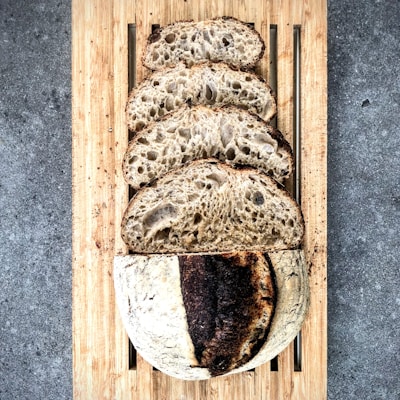 Five slices of rustic sourdough bread are placed on a wooden cutting board. The bread has a crusty exterior with a slightly burnt section and a porous, airy interior. The background features a textured, gray stone surface.