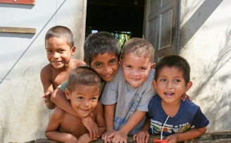 Five children are gathered closely together, smiling and leaning towards the camera. They appear to be standing in the doorway of a building with an open door behind them. They are dressed casually, and the background shows part of the exterior wall and some shadow patterns.
