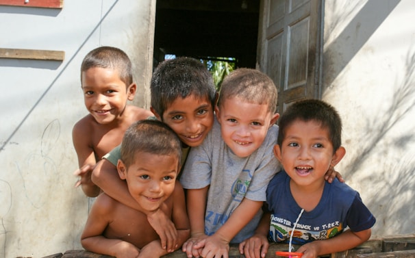 Five children are gathered closely together, smiling and leaning towards the camera. They appear to be standing in the doorway of a building with an open door behind them. They are dressed casually, and the background shows part of the exterior wall and some shadow patterns.