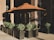 An outdoor seating area with large brown umbrellas providing shade over a few tables and chairs. The seating is surrounded by large planters containing green shrubs. The background features large stone columns and part of a building entrance, indicating it might be a café or restaurant setup.