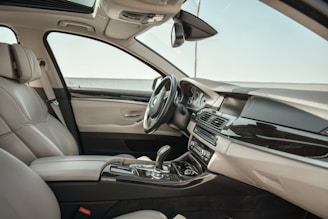 The image depicts the interior of a luxury car, featuring plush beige leather seats, a sleek dashboard with modern controls, and a shiny gear shift console. There are chrome and black accents throughout, and the car features a sunroof that allows natural light to enter the cabin.