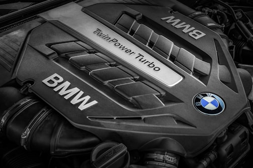 A close-up view of a BMW engine cover featuring the words 'TwinPower Turbo' and the BMW logo prominently displayed. The engine components have a sleek, metallic design with visible grooves and contours.
