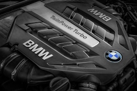 A close-up view of a BMW engine cover featuring the words 'TwinPower Turbo' and the BMW logo prominently displayed. The engine components have a sleek, metallic design with visible grooves and contours.