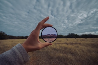 A hand holds a circular lens or filter against a backdrop of an expansive grassy field and cloudy sky. The lens magnifies and alters the appearance of a portion of the landscape, highlighting shrubs and a distant hill in warmer tones compared to the rest of the scene.