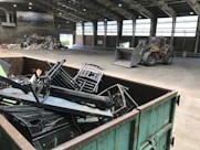 Inside a spacious industrial warehouse, a dumpster filled with discarded metal objects like chairs and other scrap is visible in the foreground. A large industrial loader is parked further inside near a pile of miscellaneous debris, including cardboard and plastic. The area is well-lit with overhead windows in the high ceiling and a poster of a landscape is mounted on the wall.