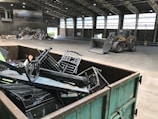 Inside a spacious industrial warehouse, a dumpster filled with discarded metal objects like chairs and other scrap is visible in the foreground. A large industrial loader is parked further inside near a pile of miscellaneous debris, including cardboard and plastic. The area is well-lit with overhead windows in the high ceiling and a poster of a landscape is mounted on the wall.