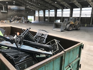 Inside a spacious industrial warehouse, a dumpster filled with discarded metal objects like chairs and other scrap is visible in the foreground. A large industrial loader is parked further inside near a pile of miscellaneous debris, including cardboard and plastic. The area is well-lit with overhead windows in the high ceiling and a poster of a landscape is mounted on the wall.