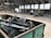 Inside a spacious industrial warehouse, a dumpster filled with discarded metal objects like chairs and other scrap is visible in the foreground. A large industrial loader is parked further inside near a pile of miscellaneous debris, including cardboard and plastic. The area is well-lit with overhead windows in the high ceiling and a poster of a landscape is mounted on the wall.