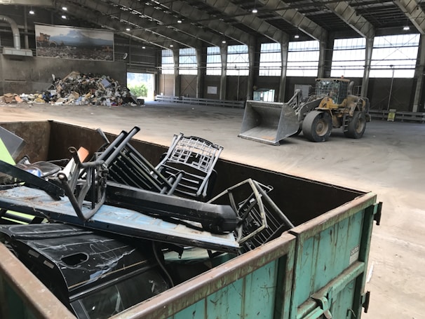 Inside a spacious industrial warehouse, a dumpster filled with discarded metal objects like chairs and other scrap is visible in the foreground. A large industrial loader is parked further inside near a pile of miscellaneous debris, including cardboard and plastic. The area is well-lit with overhead windows in the high ceiling and a poster of a landscape is mounted on the wall.