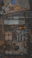 An aerial view of a construction site with visible steel frames and concrete foundations. A yellow crane or construction vehicle is present, along with scattered building materials and equipment. The overall layout suggests the initial phases of building construction.