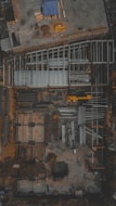 An aerial view of a construction site with visible steel frames and concrete foundations. A yellow crane or construction vehicle is present, along with scattered building materials and equipment. The overall layout suggests the initial phases of building construction.