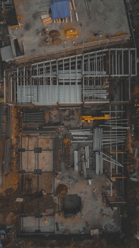 An aerial view of a construction site with visible steel frames and concrete foundations. A yellow crane or construction vehicle is present, along with scattered building materials and equipment. The overall layout suggests the initial phases of building construction.
