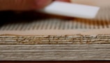A close-up view of the edge of an old, weathered book with thick, yellowed pages. There is a hand holding a white tool near the book, possibly for cleaning or preservation.