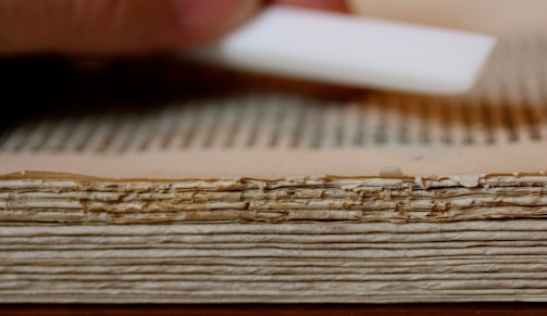 A close-up view of the edge of an old, weathered book with thick, yellowed pages. There is a hand holding a white tool near the book, possibly for cleaning or preservation.