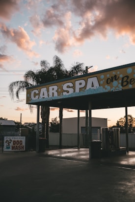 A car wash facility with a sign reading 'CAR SPA' and an additional small sign indicating a coin-operated dog wash. The scene is set at sunset or sunrise with a partly cloudy sky in the background. Tall palm trees are visible around the facility, and there is a quiet, almost deserted atmosphere.