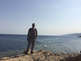 A person stands on a rocky shoreline with a vast expanse of ocean stretching out behind them. The horizon is clear, and distant mountains are visible on the right side. The sky is bright and clear, giving a sense of openness.