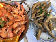 A top view of a platter filled with cooked shrimp and a dish with fried sardines. The shrimp are garnished with a few pieces of parsley and an orange slice, while the fried sardines are topped with black olives and fresh parsley. The dishes are served on a white tablecloth, partially showing silverware.