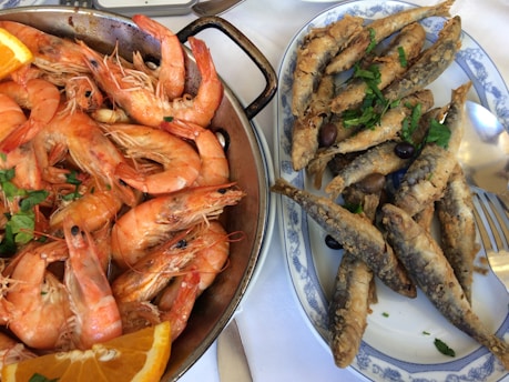 A top view of a platter filled with cooked shrimp and a dish with fried sardines. The shrimp are garnished with a few pieces of parsley and an orange slice, while the fried sardines are topped with black olives and fresh parsley. The dishes are served on a white tablecloth, partially showing silverware.
