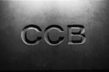 Letters 'CCB' are engraved in a concrete or stone surface, with subtle shadows creating a three-dimensional effect. The lighting is soft, emphasizing the texture of the material.