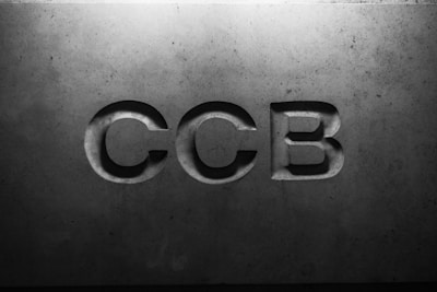 Letters 'CCB' are engraved in a concrete or stone surface, with subtle shadows creating a three-dimensional effect. The lighting is soft, emphasizing the texture of the material.