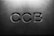 Letters 'CCB' are engraved in a concrete or stone surface, with subtle shadows creating a three-dimensional effect. The lighting is soft, emphasizing the texture of the material.