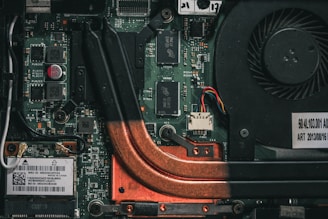 A close-up view of a computer motherboard with visible circuit paths, copper cooling pipes, electronic components, and a cooling fan. There are various chips, transistors, and capacitors mounted on the green circuit board.