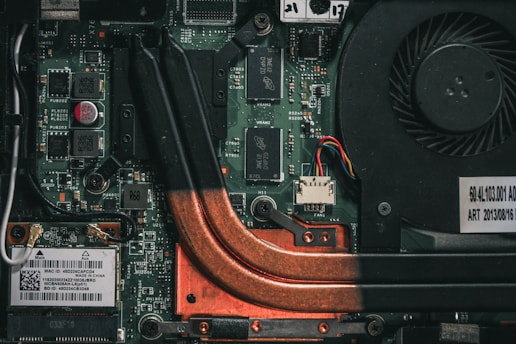 A close-up view of a computer motherboard with visible circuit paths, copper cooling pipes, electronic components, and a cooling fan. There are various chips, transistors, and capacitors mounted on the green circuit board.