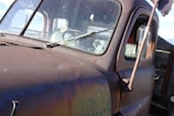 The image features a close-up view of an old, rusty vehicle, possibly a truck, with an aged and weathered exterior. The windshield and side mirrors are visible, covered in dirt and grime. The passenger seat, partially visible through the window, appears to have a worn-out, pale green seat cover. The surroundings suggest an outdoor setting.