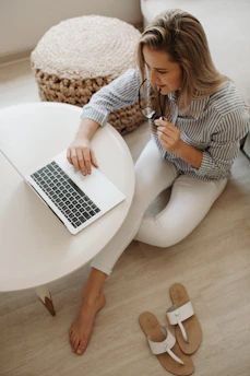 woman in white and black striped dress shirt sitting on floor in front of table while using laptop computer
